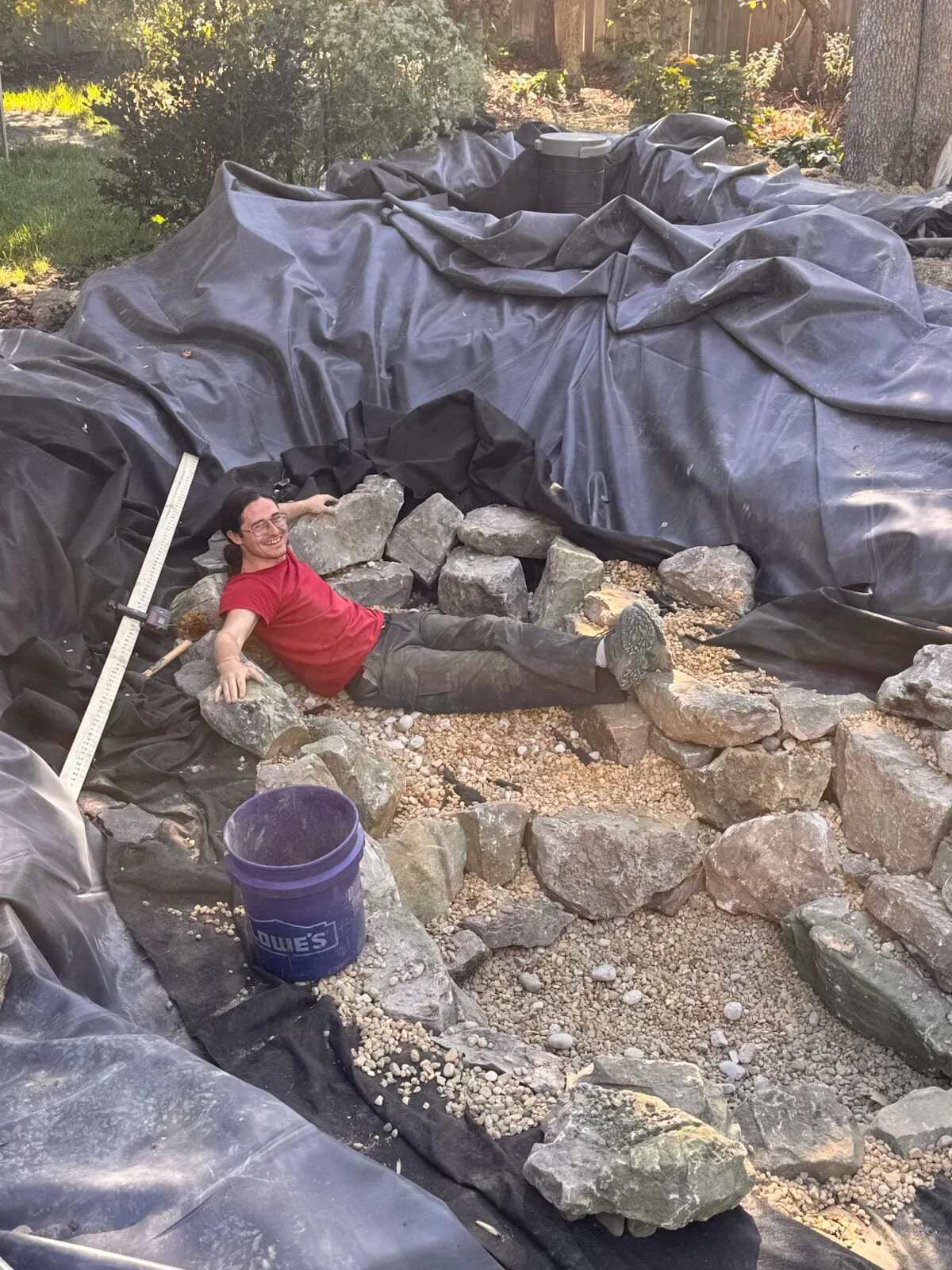 A crew member in a red shirt setting the rubber liner and first boulders in a new pond